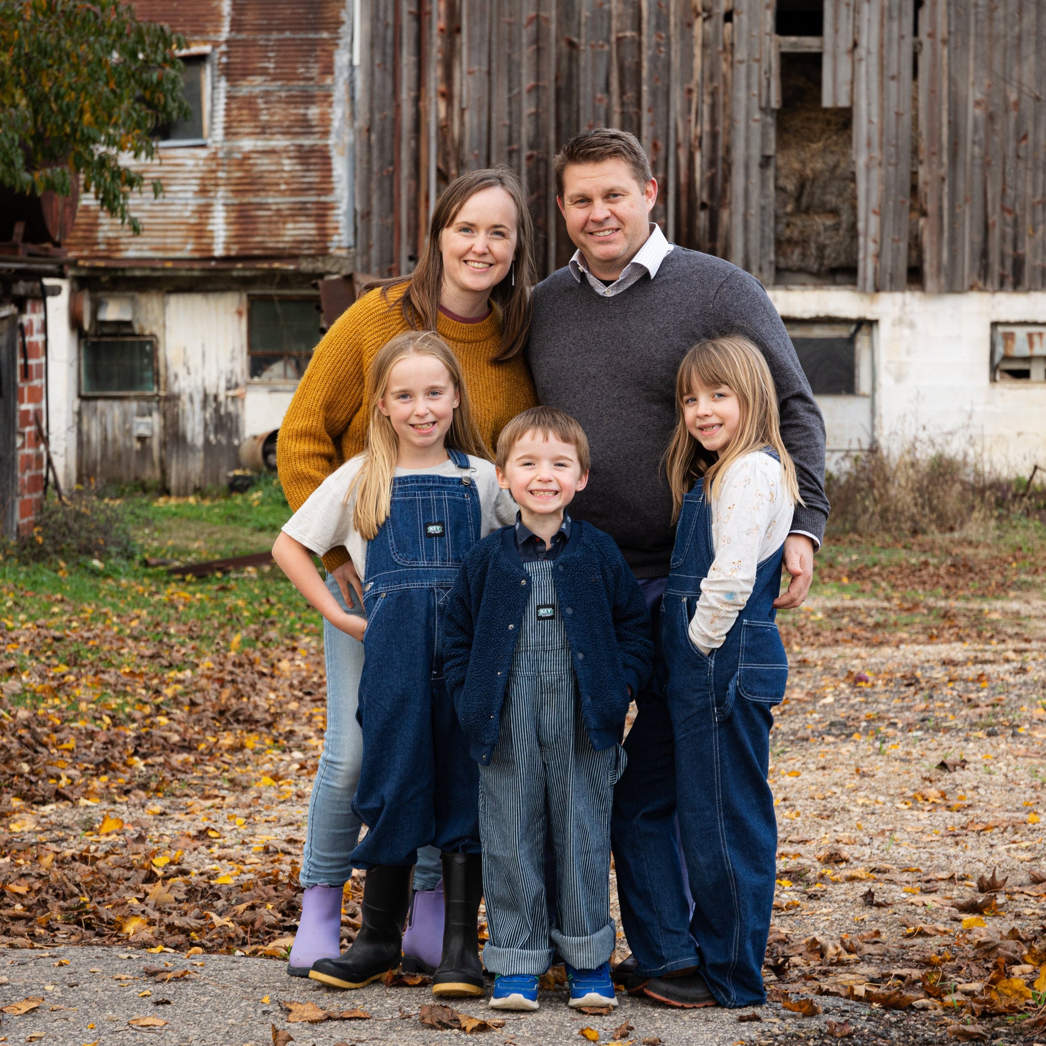 Family of five standing in front of an old barn with autumn leaves on the ground.