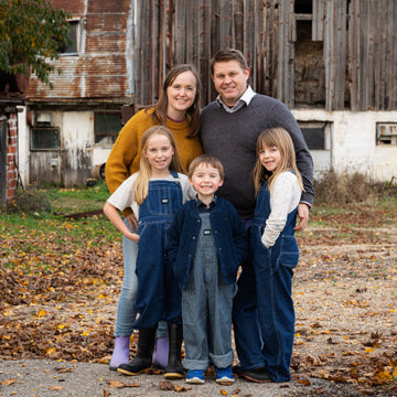 Family of five standing in front of an old barn with autumn leaves on the ground.